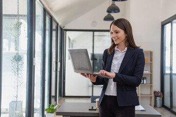 A stylish white businesswoman in a black suit standing inside the office in hands with a mobile phone, tablet, and laptop waiting for customers to contact, business people concept.