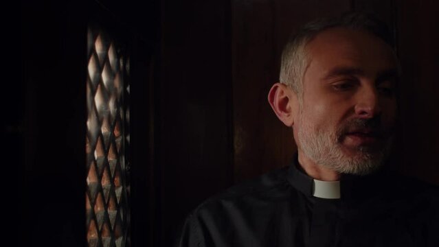 Tired middle-aged priest sitting in the confessional. The priest sighs and shakes his head negatively during confession.