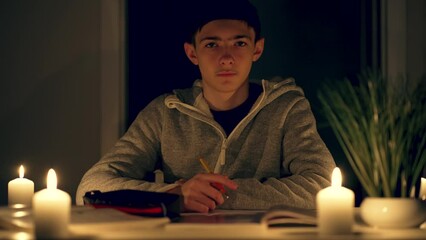 Schoolboy looking at camera while writing and teaching offline by candlelight at night. Children doing school homework at home. Studying without electricity
