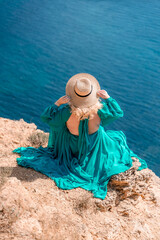 Woman sea. A happy girl is sitting with her back to the viewer in a mint dress on top of a mountain against the background of the ocean and rocks in the sea.