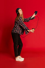 Young beautiful positive girl in checkered shirt and cap listening to music in headphones and dancing with coffee over red studio background. Concept of youth, beauty, fashion, lifestyle, emotions. Ad