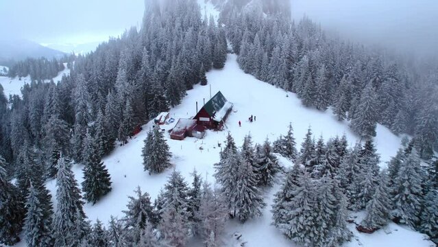 Aerial drone view of mountains in winter. Mountain peak with house and tour group nearby, firs covered with snow, low clouds