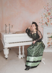 Beautiful thoughtful woman wearing green medieval vintage Victorian Style dress sitting near piano