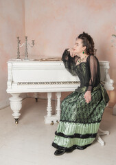 Beautiful thoughtful woman wearing green medieval vintage Victorian Style dress sitting near piano