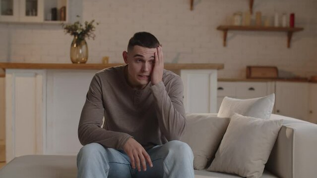 A worried man clutches his head while sitting on the couch. The short-haired man clutches his head and sighs in anticipation of bad news.
