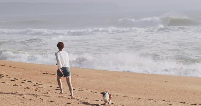 Woman running on an ocean beach sand with her dog. Female having fun with her pet cavalier king charles spaniel at Praia Norte in Nazare, Portugal. Sunny day, big waves, stormy ocean. Active vacation.