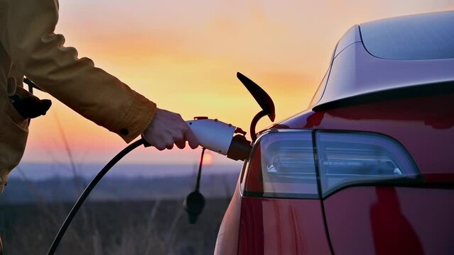 Close view of a man plugging charging cable into a parked red electric car at sunset