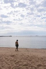 Naklejka premium Vertical photo of a young latin man walking towards the water on a sandy beach on a cloudy day. Copy space.