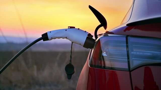 Close view of a man plugging charging cable into a parked red electric car at sunset