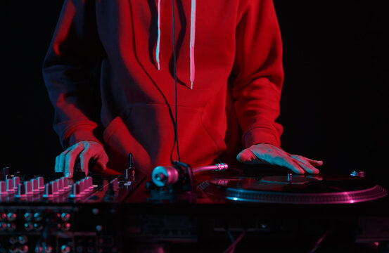 Cool young DJ in red hoodie scratching vinyl records on turntables in night club. Hands of a hip hop disc jockey playing music on stage
