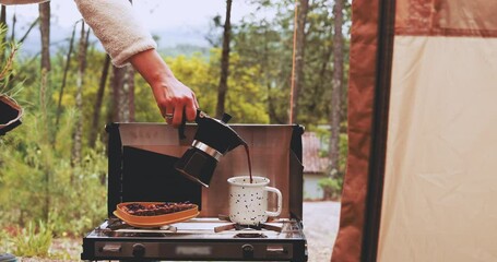 Pouring coffee in tin cup from moka pot. Food on a portable gas stove near camping tent. Peanut butter and jelly toast and freshly brewed espresso for slow breakfast in forest outdoors 