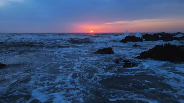 Aerial footage of stormy ocean at sunset. Flight over waves crushing on rocky stones shore at sunrise. Calm relaxing meditative tranquil nature background of morning, evening or dusk sea. 