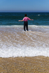 Fototapeta na wymiar A teenager girl with dark hair in a wet pink t-shirt stands knee-deep in water facing the ocean
