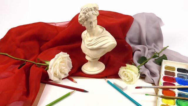 Composition for drawing from life of a gypsum cast male bust face model, watercolors and paint brushes and white fresh roses against the red textile background in an art school 