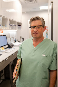 Portrait Of Male Doctor In Scrubs At Clinic Nurses Station