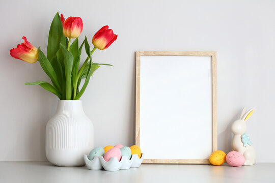 Happy Easter home decor on table on white wall background. Picture frame template, vase of red tulips, Easter eggs and bunny.