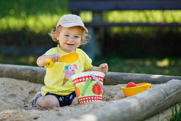 Happy toddler girl playing in sand on outdoor playground. Baby having fun on sunny warm summer...