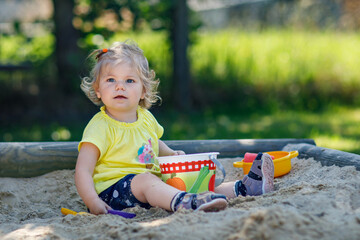 Happy toddler girl playing in sand on outdoor playground. Baby having fun on sunny warm summer sunny day. Active child with sand toys and in colorful fashion clothes.