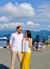 Happy couple of lovers walking at the harbour. Mountains at background. Cheerful, smiling man in white and woman in white blouse and yellow trousers, outdoors at sea port.