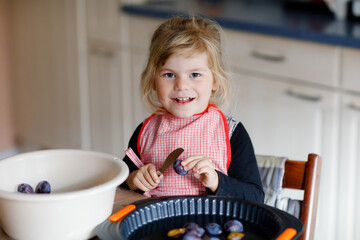 Cute little toddler girl baking plum pie at home. Happy smiling child helping and preparing plums...