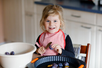 Cute little toddler girl baking plum pie at home. Happy smiling child helping and preparing plums for cake in domestic kitchen. Healthy homemade food.