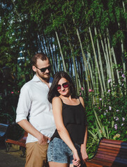 Happy couple of lovers walking outdoors. Bamboo gardens at background. Cheerful, smiling man and lucky woman in sunglasses have fun time in summer.