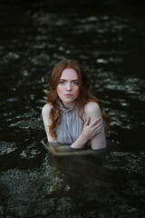 Young woman with long hair standing in water and hugging herself. Red head female adult stand in wavy river. 