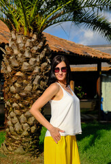 Romantic woman posing near big tree palm. Cheerful, smiling, lucky woman in white blouse, yellow trousers and sunglasses, outdoors. Sunny summer day.