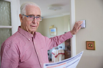 Worried Senior Man With Bill Turning Down Central Heating Thermostat At Home In Energy Crisis