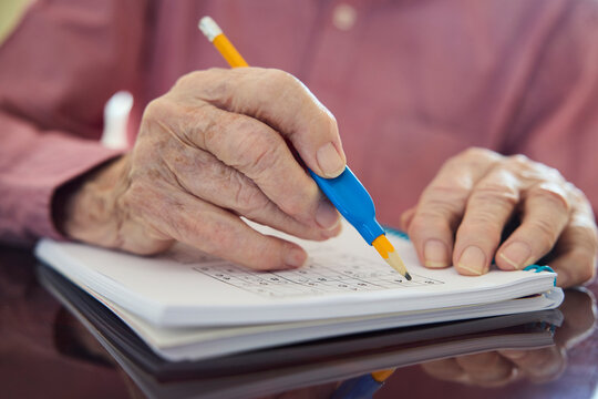 Close Up Of Senior Man With Arthritis Using Grip Aid On Pencil To Write In Sudoku Puzzle Book