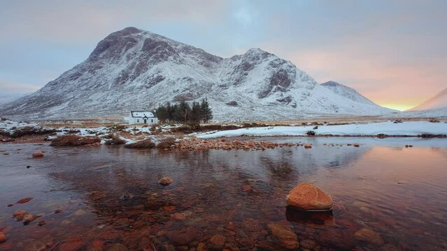 A remote hut and calm river with stone on the foreground at the foot of Buachaille Etive Mor at the entrance to the valley of Glencoe in the Scottish Highlands, Scotland