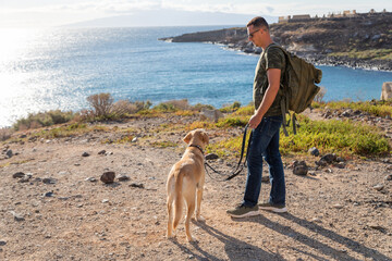 A traveler wears his backpack and keeps his dog on a leash near the sea