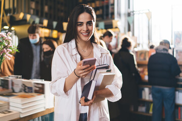 Cheerful woman standing with smartphone and books in bookshop