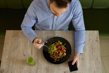 Mid adult man enjoys eating salad, drinking smoothie and surfing the net on smart phone in salad bar