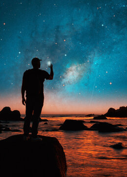 silhouette of a person on a rock taking pictures with his cell phone at night of the milky way on the horizon over the ocean