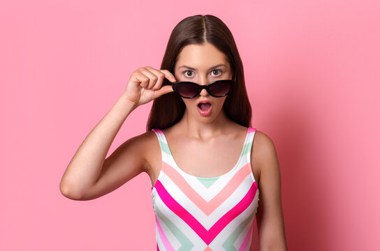 amazed surprised teen girl with open mouth taking off sunglasses expressing surprise, dressed in colourful one-piece swimsuit isolated on pink background, summer vacation holidays