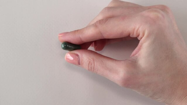 Dark green capsule with chlorophyll or spirulina in a woman's hand. Helpful supplements or medicines on a beige background.