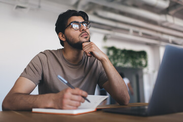 Thoughtful man taking notes while sitting with laptop