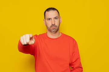 Bearded Hispanic man in a red sweater pointing at the camera with his finger, he is very serious and has an accusatory attitude. Isolated on yellow studio background.