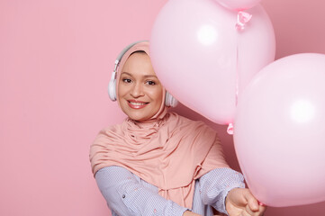 Beautiful Middle-Eastern Muslim woman wearing wireless headphones and pink hijab, holding out at camera a bunch of pastel helium party balloons, smiling looking at camera, isolated color background