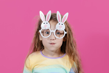 Cute little child girl wearing bunny ears glasses a on Easter day. Easter girl portrait on pink background, funny emotions, surprise.