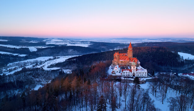 Aerial view of romantic, fairy castle in winter highland covered in snow, illuminated by pink light. Gothic castle in picturesque landscape, high towers, red roofs,  winter season. Bouzov, Czechia.