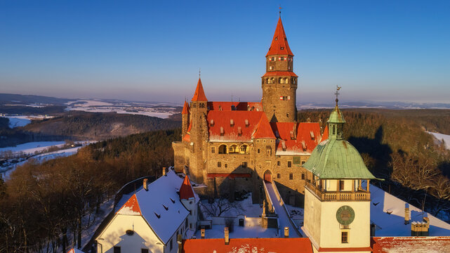 Aerial view of romantic, fairy castle in winter picturesque landscape, illuminated by the setting sun. Gothic castle, high towers, red roofs,  winter highland covered in snow. Bouzov castle, Czechia. 
