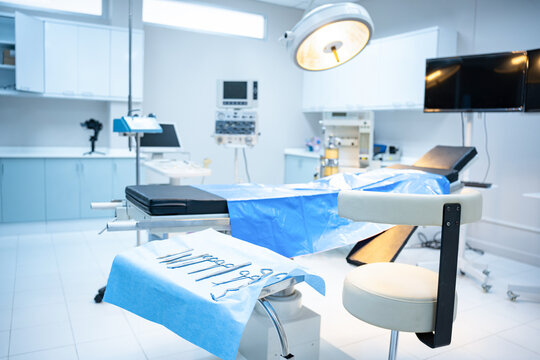 Interior of modern operating room with selective focus of empty doctor's chair beside surgical instruments set on blue cloth, with an empty operating bed with surgical light shining down in background