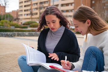 Portrait of two young woman students in university during break, carrying laptop and books in hand,...