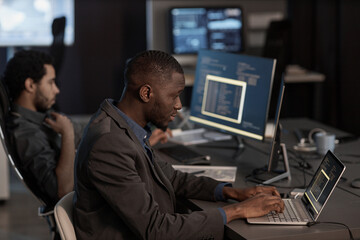 Side view portrait of black man writing code and typing at laptop keyboard while working in data...