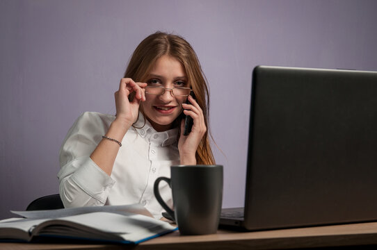 The girl is sitting at a desktop with a computer and talking on a mobile phone.
