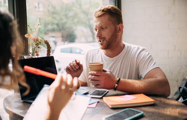 Skilled male and female discussing program code and web design for IT startup project during collaboration job in cafeteria interior, Caucaisan freelancers with laptop and paperwork communicate