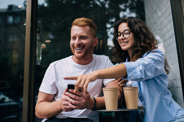 Joyful male blogger with smartphone and female in optical eyewear smiling and discussing leisure during coffee break at street cafeteria, happy Caucaisan couple in love enjoying friendly relations