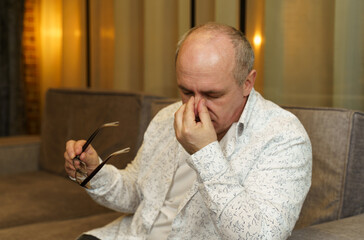 A mature man holds glasses with his hand, the other rubs his hands near his eyes.